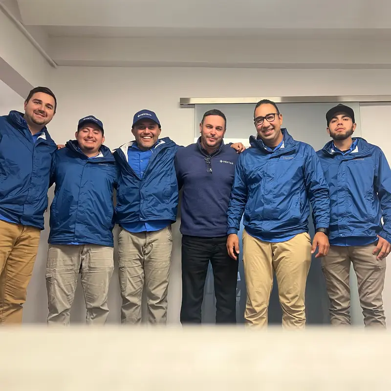 Group of six men standing indoors wearing blue jackets and khaki pants smiling at the camera.