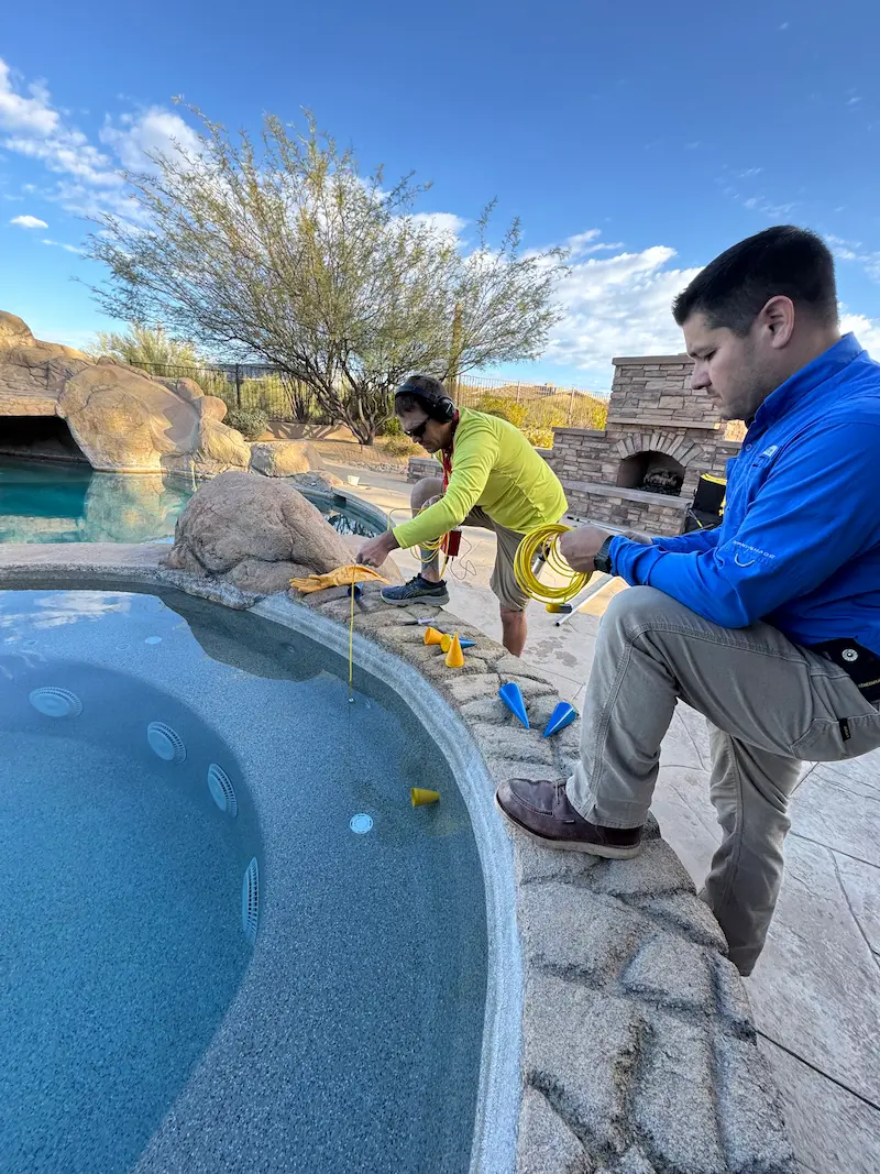 Two men testing water quality in a backyard pool and spa area using measuring tools.